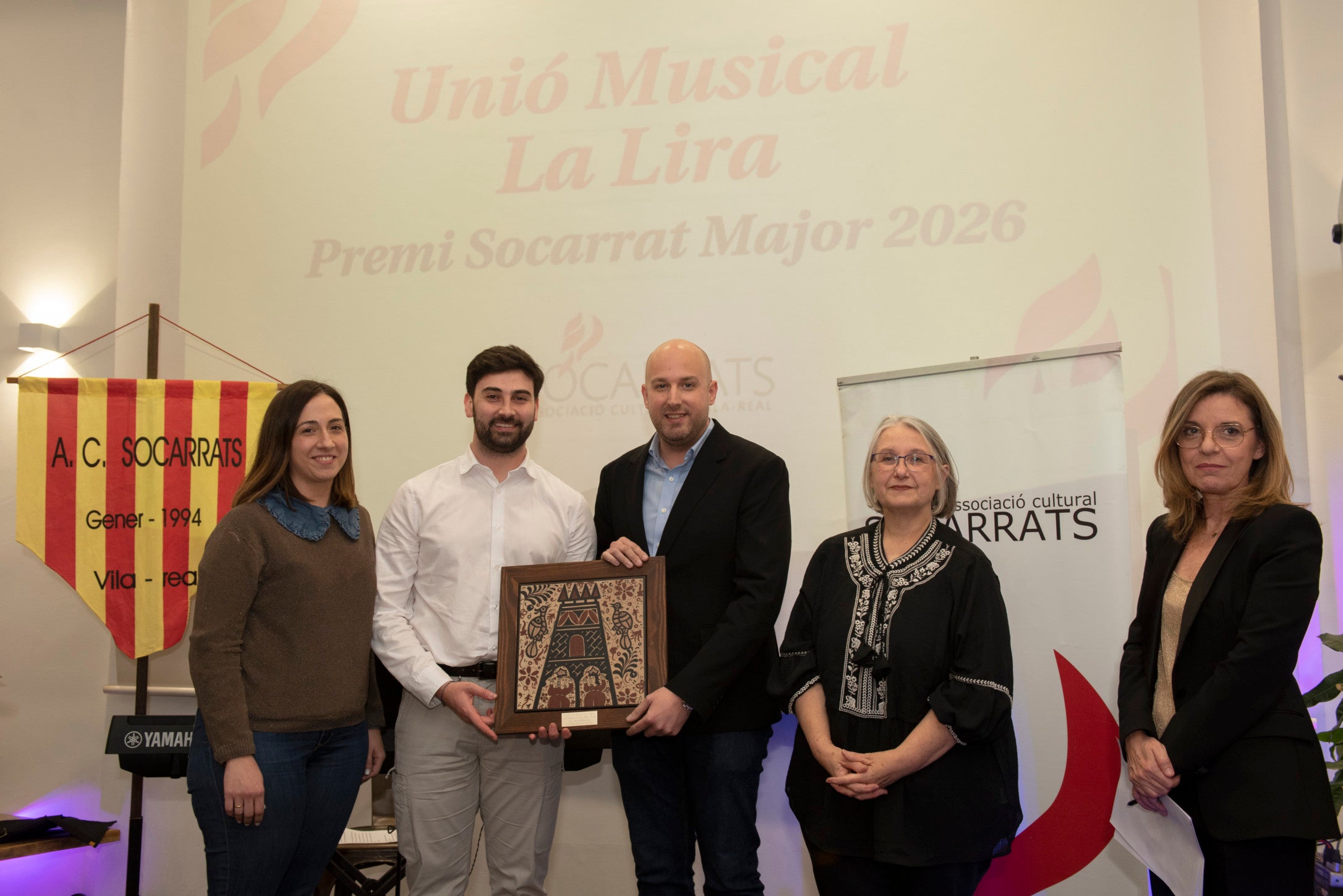 El vicepresident de la Unió Musical La Lira, David Cubedo, acompanyat d’altres membres de la Junta, Pilar Marí i Manel Boixader, rep el premi Socarrat Major 2026 